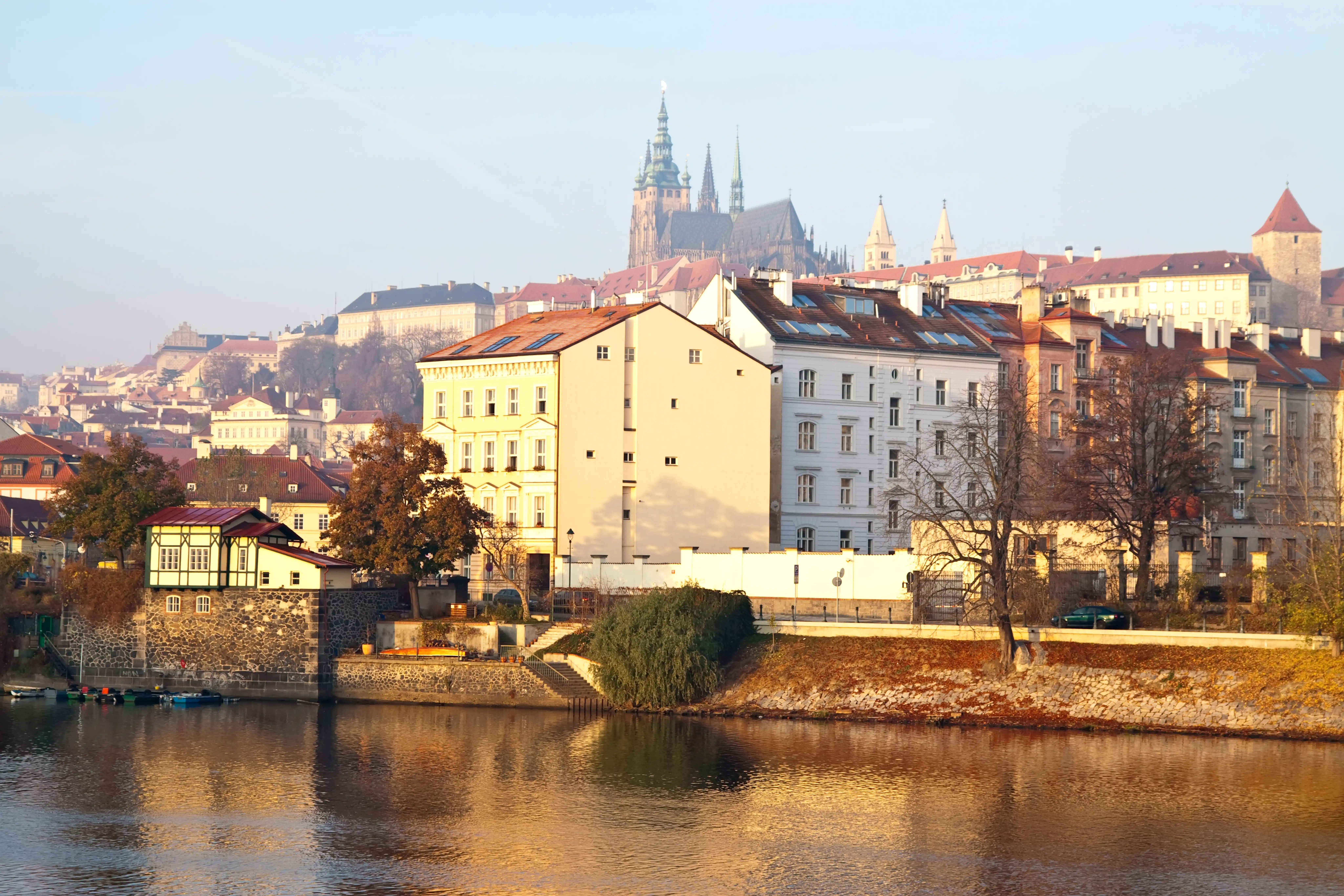 Historic buildings along a riverbank with a large cathedral and towers in the background under a clear sky.