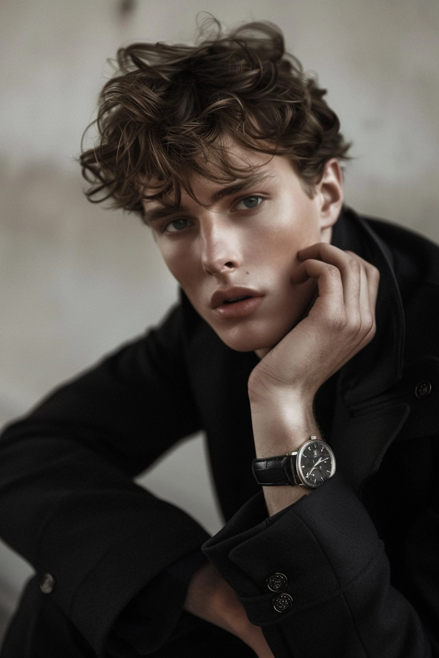 Close-up portrait of a young man with wavy brown hair wearing a black coat and wristwatch.
